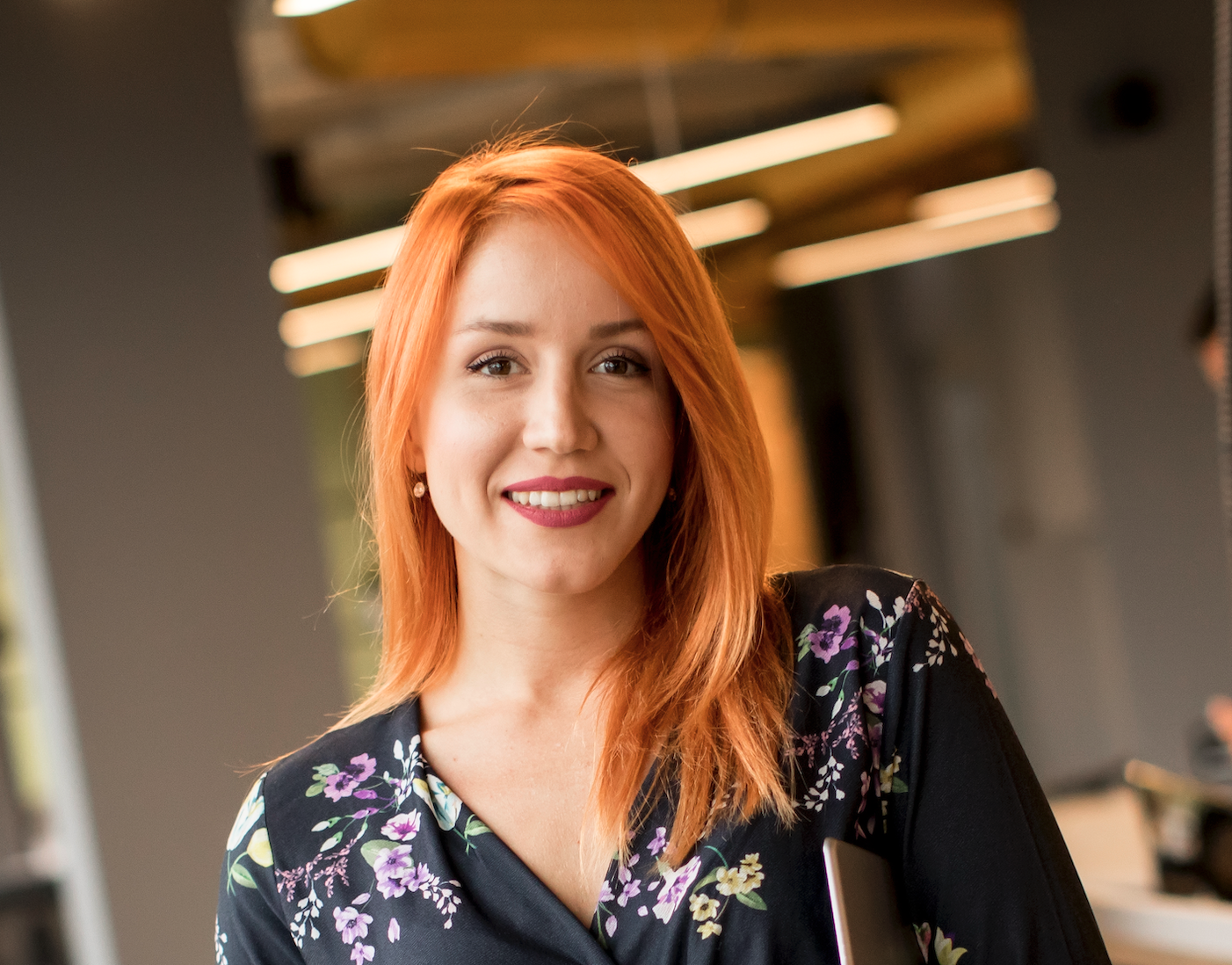Smiling woman with orange hair wearing a floral blouse, holding a laptop, in a modern office setting.